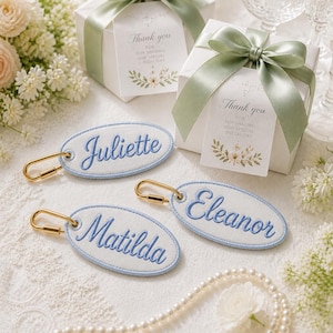 May include: Three oval white fabric tags with light blue embroidered borders and names Juliette, Eleanor, and Matilda in blue script. Each tag features a gold-toned metal lobster clasp. Two white gift boxes with sage green ribbons are present.