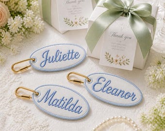 Könnte beinhalten: Drei ovale weiße Stoffanhänger mit hellblau bestickten Rändern und den Namen Juliette, Eleanor und Matilda in blauer Schreibschrift. Jeder Anhänger hat einen goldfarbenen Karabinerverschluss aus Metall. Zwei weiße Geschenkboxen mit salbeigrünen Bändern.