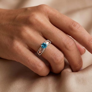 May include: A silver ring with a round, bright blue gemstone. The ring has a leaf design with rose gold accents and small, clear stones. The ring is on a finger, with a soft, neutral background.