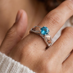May include: A silver ring with a large, round, bright blue gemstone. The ring has a decorative design with gold-colored leaf accents on the sides. The ring is on a finger.
