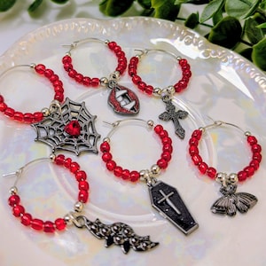 May include: A collection of wine charms with red beaded hoops and various charms. Charms include a spiderweb with a red gem, a coffin, a cross, a bat, a pair of fangs, and a moth. The charms are displayed on a white plate.