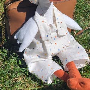 May include: A handmade goose plush toy wearing a white outfit with colorful confetti-like patterns. The goose has a white head, orange beak and feet, and is resting on a brown purse on a bed of green grass.