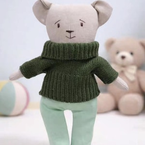 May include: A handmade stuffed animal mouse wearing a dark green knit sweater and light green pants. The mouse has a light beige head and ears, with embroidered facial features. A second teddy bear is visible in the background.