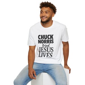 May include: White t-shirt with the text "CHUCK NORRIS tried JESUS LIVES" in black block letters. The shirt is short-sleeved and worn by a person wearing blue jeans.