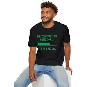 Peut inclure: T-shirt noir avec le texte "ENLIGHTENMENT PENDING... PLEASE HOLD_" en vert. Le t-shirt présente un graphique de barre de chargement. Le modèle porte un jean bleu.