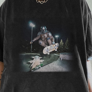 May include: A charcoal gray oversized t-shirt featuring a graphic print of a character in armor skateboarding in the air above a small creature on a skateboard. The shirt has short sleeves, a crew neckline, and a faded, washed-out fabric texture.