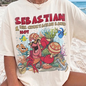 以下が含まれることがあります： オフホワイトの半袖Tシャツ。マラカスを演奏するカニのセバスチャンのグラフィックプリントが施され、上部には「SEBASTIAN & HIS CRUSTACEAN BAND HOT」の文字があり、周囲には海中のサンゴ、魚、錨のイラストが描かれています。