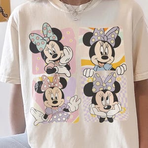 May include: Cream-colored t-shirt featuring a Minnie Mouse graphic. The graphic showcases four different Minnie Mouse poses, each within a pastel-colored square. Minnie is wearing polka-dot bows and dresses. The shirt has short sleeves and a crew neck.