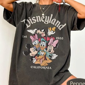 May include: A dark gray t-shirt featuring the word "Disneyland" above a graphic of Disney characters, including Mickey and Minnie Mouse, Donald Duck, and Goofy. The shirt also includes the text "Est 1955" and "California".