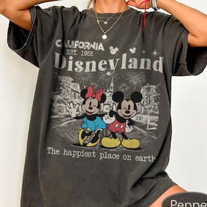 May include: Dark gray t-shirt featuring a graphic of Mickey and Minnie Mouse in front of Disneyland. The shirt has the text "CALIFORNIA EST. 1955 Disneyland" and "The happiest place on earth."