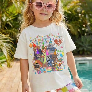 May include: A young girl wearing a white t-shirt with a colorful graphic of cartoon animals and the text "HAPPY BIRTHDAY 3". She is also wearing pink flower-shaped sunglasses, a purple headband, and checkered shorts.