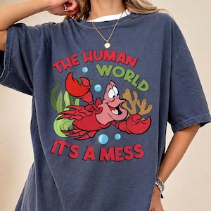 May include: A navy blue t-shirt featuring a cartoon image of a red crab with the text "THE HUMAN WORLD IT'S A MESS" in red and green. The shirt has a relaxed fit and a white undershirt is visible.