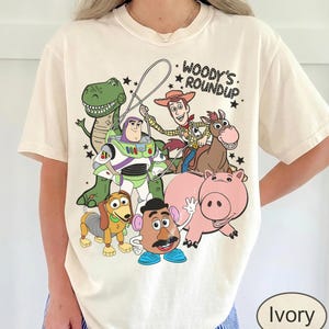May include: Ivory-colored t-shirt featuring a graphic of characters from the animated film "Toy Story." The design includes Woody, Buzz Lightyear, Rex, Hamm, Slinky Dog, Mr. Potato Head, and a horse. Text above the characters reads "Woody's Roundup."