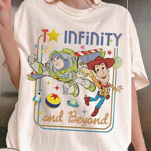 May include: Ivory-colored t-shirt featuring a graphic of Buzz Lightyear and Woody from Toy Story. The design includes the text "To Infinity and Beyond" with stars and other playful elements. The shirt has a crew neck and short sleeves.