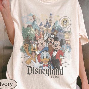 May include: An ivory short-sleeve t-shirt featuring a graphic print of Mickey Mouse, Minnie Mouse, Donald Duck, Goofy, and Pluto in front of a Disneyland castle. The text "Disneyland Est. 1955" is printed in a dark font below the character illustration.