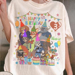 May include: Ivory-colored t-shirt featuring a colorful birthday design with cartoon characters, balloons, and the text "Happy Birthday." The number "3" is displayed, along with space for a name. Party hats and gifts are also included.