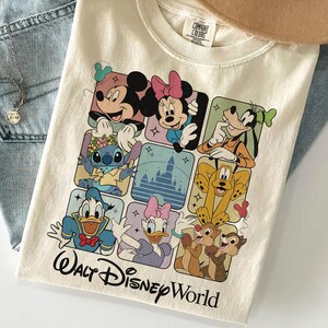 May include: Cream-colored t-shirt featuring a grid of Disney characters including Mickey, Minnie, Goofy, Stitch, Donald Duck, Daisy, Chip, and Dale. The words "Walt Disney World" are printed in black script at the bottom.