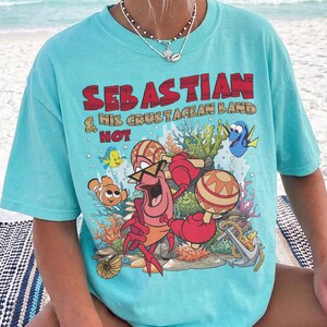 以下が含まれることがあります： 明るいターコイズブルーのTシャツ。セバスチャンというカニが他の海の生き物たちとマラカスを演奏しているグラフィックがプリントされています。テキストは「SEBASTIAN & HIS CRUSTACEAN BAND HOT」と書かれています。デザインにはサンゴ、錨、泡が含まれています。