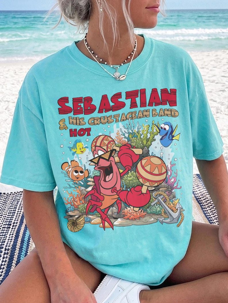 以下が含まれることがあります： 明るいターコイズブルーのTシャツ。セバスチャンというカニが他の海の生き物たちとマラカスを演奏しているグラフィックがプリントされています。テキストは「SEBASTIAN & HIS CRUSTACEAN BAND HOT」と書かれています。デザインにはサンゴ、錨、泡が含まれています。