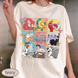 May include: Ivory-coloured t-shirt with a colourful grid design featuring characters from Disney's The Little Mermaid. The design includes Ariel, Flounder, Sebastian, King Triton, Ursula, Prince Eric, and Max. The words "The Little Mermaid" are also featured.