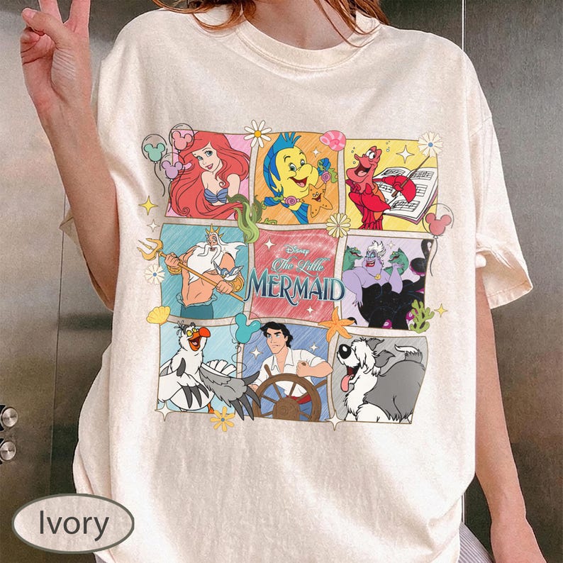 May include: Ivory-coloured t-shirt with a colourful grid design featuring characters from Disney's The Little Mermaid. The design includes Ariel, Flounder, Sebastian, King Triton, Ursula, Prince Eric, and Max. The words "The Little Mermaid" are also featured.