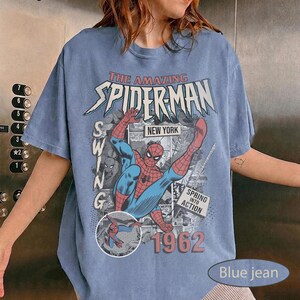 May include: Blue jean colored t-shirt featuring a graphic of Spider-Man in action. The design includes the text "THE AMAZING SPIDER-MAN", "NEW YORK", "SWING", "SPRING INTO ACTION", and the year "1962".