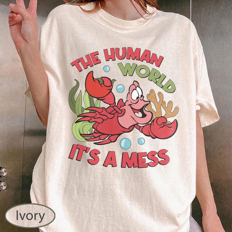 May include: Ivory-colored t-shirt featuring a cartoon crab with the text "THE HUMAN WORLD IT'S A MESS". The crab is red and pink, with green seaweed and blue bubbles. The shirt is a relaxed fit.