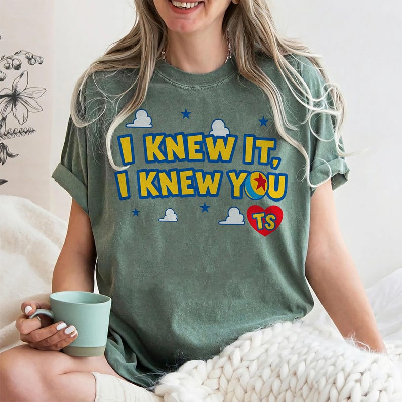 May include: A sage green short-sleeve t-shirt featuring the yellow text I KNEW IT, I KNEW YOU with a red heart containing the letters TS. The design includes small blue stars and white cloud graphics printed on the front of the garment.