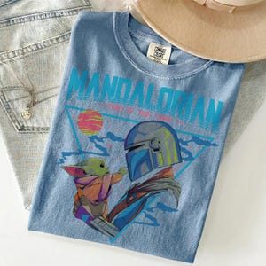 Pu&ograve; includere: Una t-shirt azzurra con una grafica del Mandaloriano e Grogu. Il testo recita "MANDALORIAN" e "THIS IS THE WAY". Il design include un pianeta rosa e il contorno di un triangolo blu. La t-shirt presenta un'etichetta del marchio Comfort Colors.