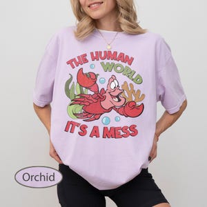 May include: Orchid-colored t-shirt featuring a cartoon image of a red crab with the text "THE HUMAN WORLD IT'S A MESS". The crab is surrounded by green seaweed and blue bubbles. The shirt is a light purple color.