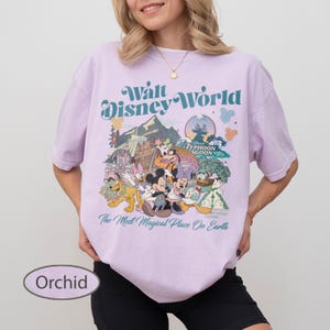 May include: Orchid-colored t-shirt featuring a vintage-style Walt Disney World graphic. The design includes various Disney characters and the text "The Most Magical Place On Earth". The shirt has short sleeves and a crew neck.