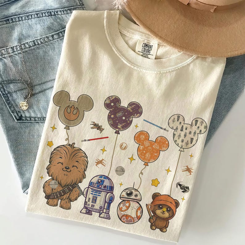 May include: Cream-colored t-shirt featuring Star Wars characters and Mickey Mouse-shaped balloons. Chewbacca, R2-D2, BB-8, and an Ewok are depicted. The balloons are in various colors and patterns, with stars and lightsabers.