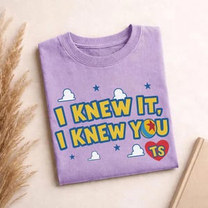 May include: A light purple crew neck t-shirt featuring the yellow text I KNEW IT, I KNEW YOU with a blue outline. The design includes a red heart with the letters TS, a colorful ball with a red star, white cloud shapes, and small blue stars.