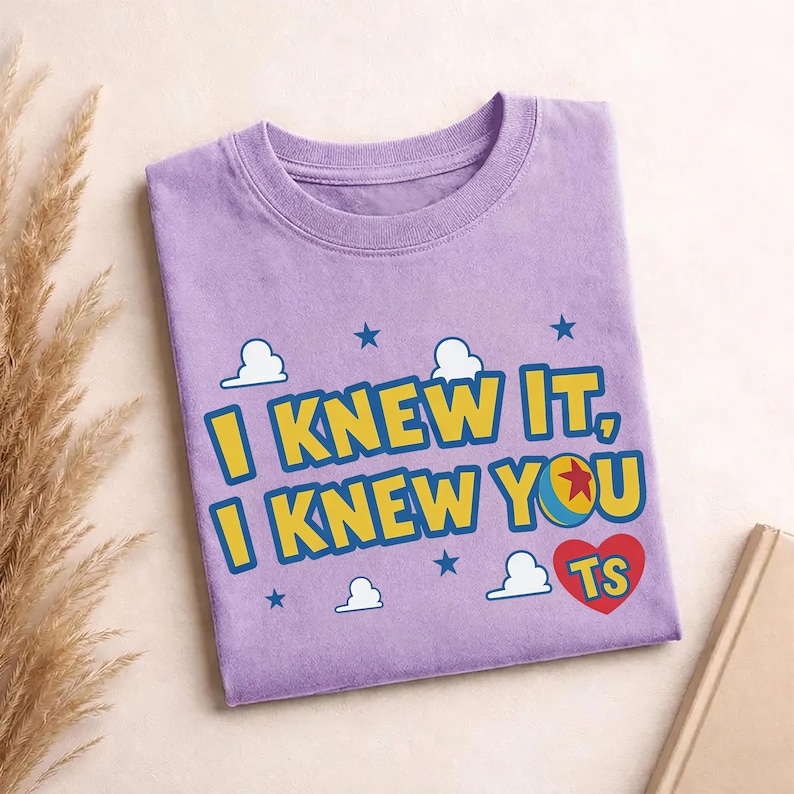 May include: A light purple crew neck t-shirt featuring the yellow text I KNEW IT, I KNEW YOU with a blue outline. The design includes a red heart with the letters TS, a colorful ball with a red star, white cloud shapes, and small blue stars.