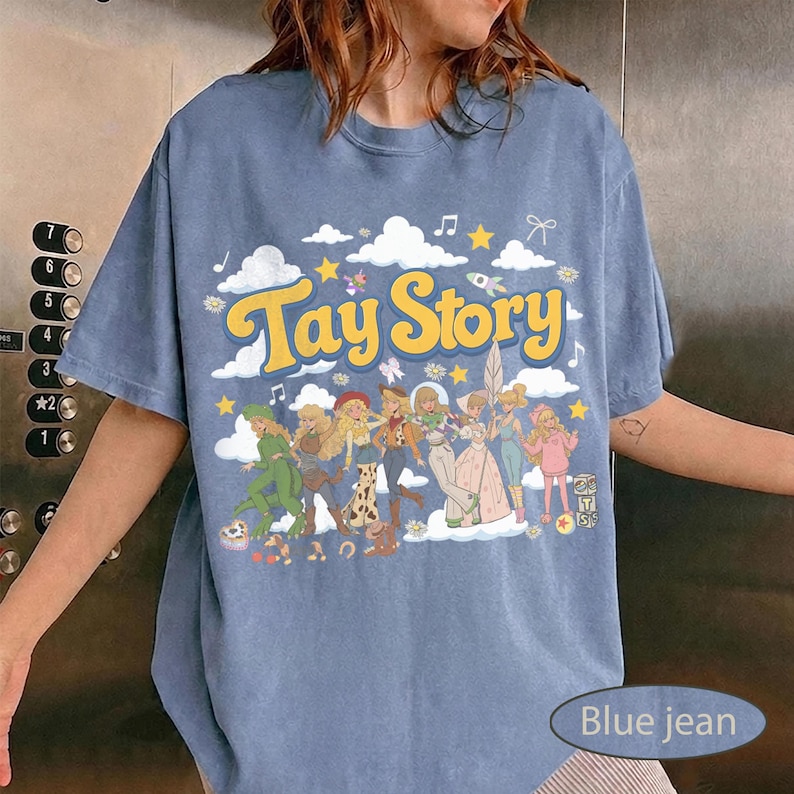 K&ouml;nnte beinhalten: Ein blaues T-Shirt in Jeansoptik mit einem Grafikdruck, der den Text Tay Story in gelben Buchstaben zeigt. Das Design enth&auml;lt Illustrationen verschiedener Figuren in unterschiedlichen Outfits, umgeben von wei&szlig;en Wolken, gelben Sternen und Noten.