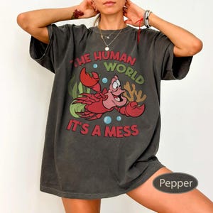 May include: Dark gray t-shirt featuring a cartoon image of a red crab with the text "THE HUMAN WORLD IT'S A MESS". The crab is surrounded by green seaweed and blue bubbles. The shirt is a relaxed fit.