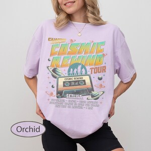 K&ouml;nnte beinhalten: Orchideenfarbenes T-Shirt mit Retro-Grafikdesign. Das Design zeigt den Text "COSMIC REWIND TOUR" mit einer Kassettenband-Illustration, Planeten und dem Text "SETLIST". Das Shirt hat eine lockere Passform.