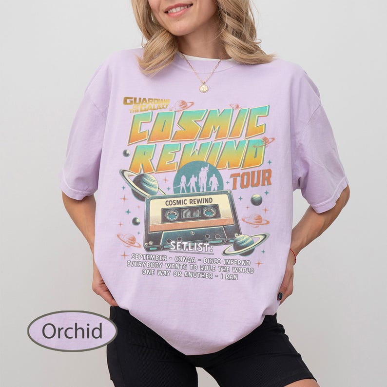 K&ouml;nnte beinhalten: Orchideenfarbenes T-Shirt mit Retro-Grafikdesign. Das Design zeigt den Text "COSMIC REWIND TOUR" mit einer Kassettenband-Illustration, Planeten und dem Text "SETLIST". Das Shirt hat eine lockere Passform.
