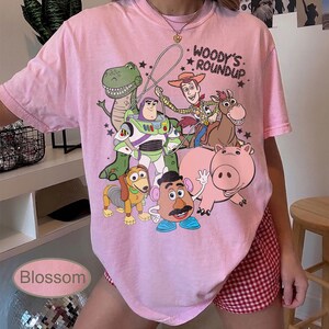 May include: Pink t-shirt featuring a colorful graphic of characters from the animated film "Toy Story." The design includes Woody, Buzz Lightyear, Rex, Hamm, Slinky Dog, and Mr. Potato Head. The text "Woody's Roundup" is also visible.