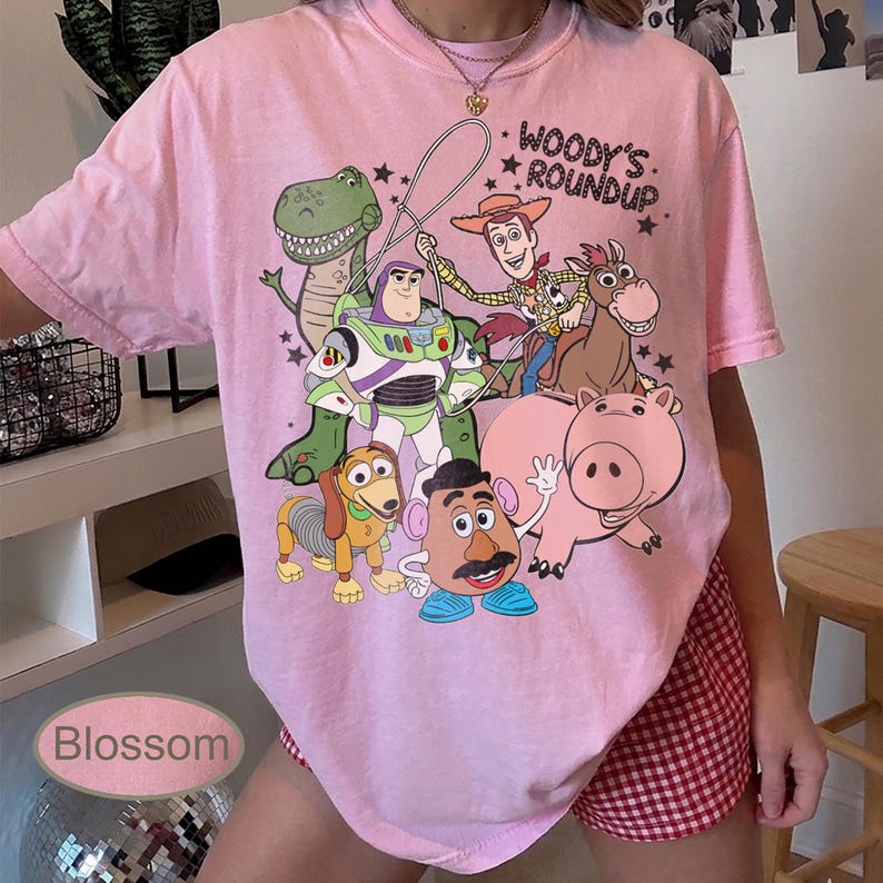 May include: Pink t-shirt featuring a colorful graphic of characters from the animated film "Toy Story." The design includes Woody, Buzz Lightyear, Rex, Hamm, Slinky Dog, and Mr. Potato Head. The text "Woody's Roundup" is also visible.