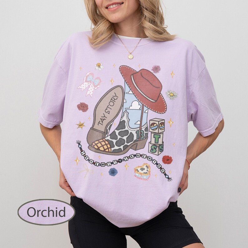 May include: A light purple t-shirt featuring a graphic print of a cow-print cowboy boot, a red cowboy hat, and building blocks with the letters T, S, and the number 13. The design includes a beaded string with text and small floral accents.