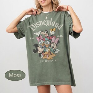 May include: Moss green t-shirt featuring the text "Disneyland" above a graphic of Disney characters. The shirt includes the text "Est 1955 CALIFORNIA". The shirt is a relaxed fit.