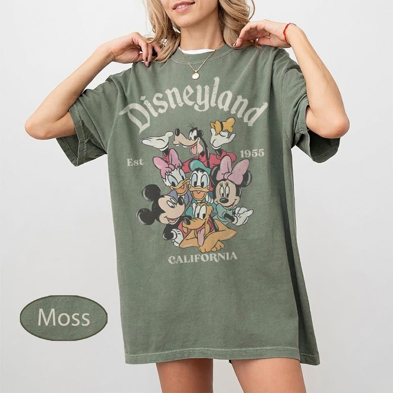 May include: Moss green t-shirt featuring the text "Disneyland" above a graphic of Disney characters. The shirt includes the text "Est 1955 CALIFORNIA". The shirt is a relaxed fit.