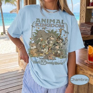 May include: Light blue t-shirt featuring a graphic of Disney characters in a jungle setting, with the text "ANIMAL KINGDOM" and "Walt Disney World". The design includes an elephant, giraffe, zebra, and other animals.