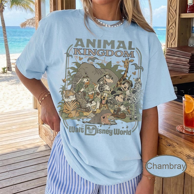 May include: Light blue t-shirt featuring a graphic of Disney characters in a jungle setting, with the text "ANIMAL KINGDOM" and "Walt Disney World". The design includes an elephant, giraffe, zebra, and other animals.