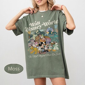 May include: Moss green t-shirt featuring a Walt Disney World graphic with Mickey Mouse, Donald Duck, and other characters. The shirt has the text "The Most Magical Place On Earth".