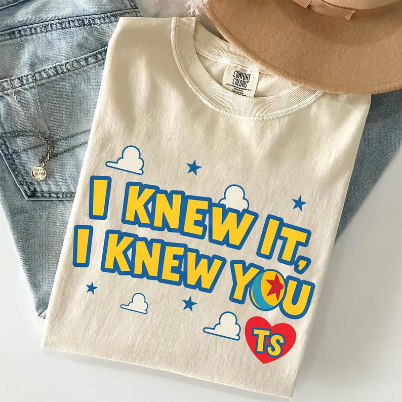 May include: A cream-colored short-sleeve t-shirt featuring the blue text I KNEW IT, I KNEW YOU with a colorful ball icon, small blue stars, white cloud outlines, and a red heart containing the yellow letters TS printed on the front chest.