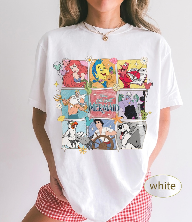 May include: White t-shirt with a colourful graphic of characters from Disney's The Little Mermaid. The design includes Ariel, Flounder, Sebastian, and other characters in a comic book style. The text "The Little Mermaid" is also present.