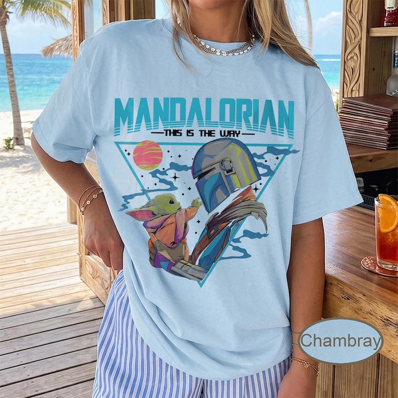 Pu&ograve; includere: Una t-shirt azzurro chiaro con una stampa grafica di The Mandalorian e Grogu. Il design include il testo "MANDALORIAN" e "THIS IS THE WAY" sopra una cornice triangolare contenente un pianeta, stelle e i due personaggi in un'illustrazione.