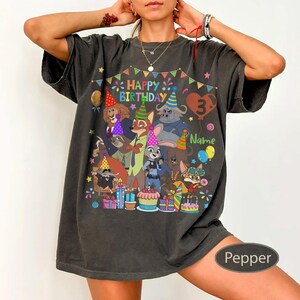 May include: A dark gray t-shirt featuring a colorful graphic of cartoon characters, party hats, and the text "Happy Birthday 3 Name". The design includes balloons, presents, and a cake. The shirt is a relaxed fit.